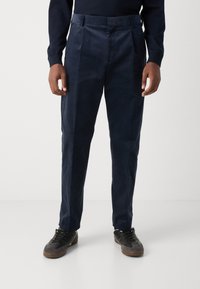 Pantalons en velours côtelé bleu marine avec une texture lisse, présentant une taille haute, des plis et des jambes droites. Associés à des baskets noires.