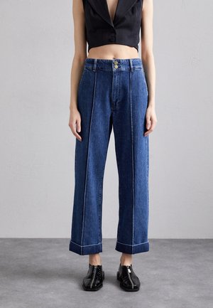 Femme portant un jean taille haute bleu avec des coutures sur le devant et de larges revers, associé à un gilet noir court et des chaussures noires brillantes à fermeture éclair.