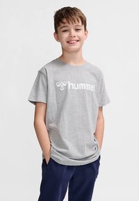 Grå bomulls-T-shirt med korta ärmar, med en vit logotyp på bröstet. Modellen bär marinblå byxor och står med händerna i fickorna.