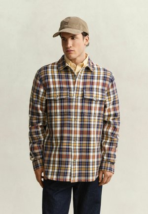 Man wearing a beige cap, yellow shirt, and a buttoned plaid jacket in blue, red, and yellow tones with dark pants, standing against a plain background.