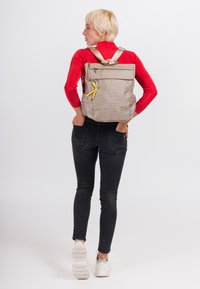 Šviesiai pilkas audinio kuprinės modelis su sukamo viršaus dizainu, reguliuojamomis juostomis ir priekinėmis kišenėmis. Sukurtas su geltonais akcentais ir tekstūruotu paviršiumi.