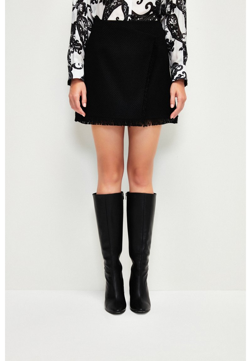Femme vêtue d’une mini-jupe noire texturée à ourlet à franges, d’un chemisier à motif cachemire et de bottes en cuir noires hautes jusqu’au genou, debout contre un mur blanc.