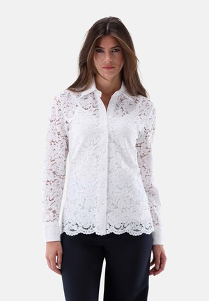 Weiße Spitzenbluse mit floralem Muster, langen Ärmeln, Knopfleiste vorne und gewelltem Saum. Der Stoff ist durchsichtig und hat eine weiche Textur.
