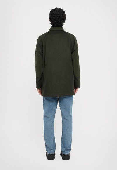 Dark green outerwear jacket with shoulder patches, loose fit, standing collar, and an open back. Paired with light blue jeans and black shoes.