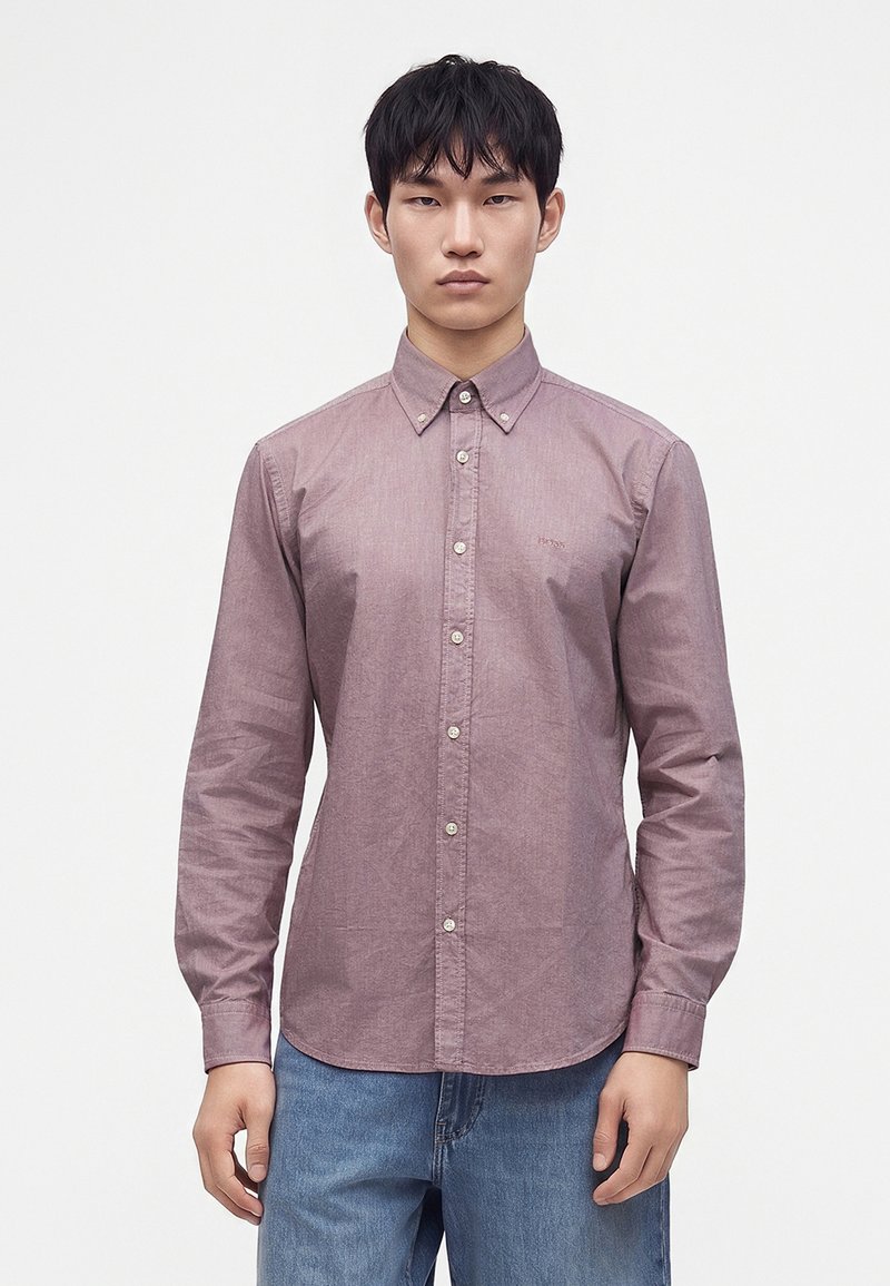 Jeune homme aux cheveux noirs courts, portant une chemise mauve à manches longues boutonnée et un jean bleu, debout devant un fond blanc uni.