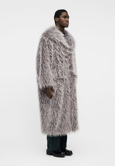 Homme portant un long manteau de fourrure gris clair et ébouriffé, un pantalon foncé et des chaussures noires, debout devant un fond clair uni, regardant vers la droite.