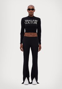 Top de malha cropped preto com o logotipo "Versace Jeans Couture", combinado com calças pretas com fendas na bainha e ténis brancos.