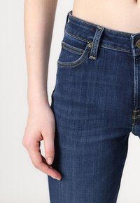 Jeans de mezclilla en azul oscuro con un acabado suave. Las características incluyen costuras doradas, cuatro bolsillos y un cierre frontal con botón.