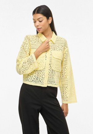Kobieta ubrana w żółtą bluzkę z długim rękawem o koronkowym wzorze z kieszeniami z przodu oraz czarne spodnie, stojąca na jednolitym białym tle.