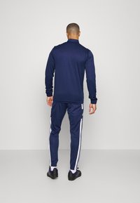 Marineblauw sporttracksuit, bestaande uit een langmouwtop en slim-fit broek met witte zijstrepen, gemaakt van een gladde stof met een lichte glans.