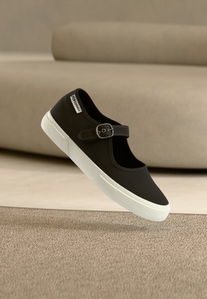 Pantof sport Mary Jane negru cu talpă de cauciuc albă și baretă reglabilă cu cataramă, plutind deasupra unui covor bej cu canapea în fundal.