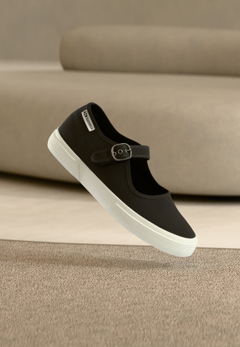 Μαύρο αθλητικό παπούτσι Mary Jane με λευκή λαστιχένια σόλα και ρυθμιζόμενο λουράκι με πόρπη, που αιωρείται πάνω από μπεζ χαλί με καναπέ στο φόντο.