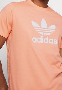 Ljus persikorosa bomulls-t-shirt med en framträdande vit Adidas-logotyp och tre horisontella ränder i designen. Kortärmad, rund halsringning.