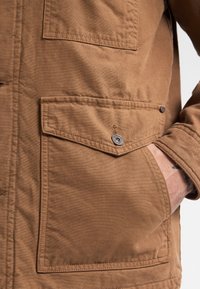 Brown canvas jacket featuring a buttoned upper pocket and a side hand pocket. The fabric is textured, with visible stitching accents.