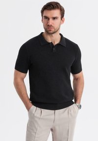 Sort kortærmet polo-shirt med et tekstureret strikmønster, en krave-udskæring og en tilpasset talje. Båret med lyse bukser.