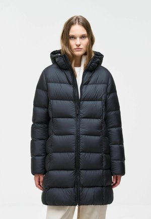 Young woman wearing a long black puffer jacket with a hood, standing against a plain light background, facing forward.