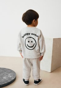 Harmaa huppari ja collegehousut, joissa takana musta "happy crew" -teksti ja hymyilevä kasvojen grafiikka. Puuvillasekoituskangas, rento malli.