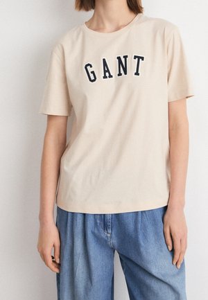 Beige Baumwoll-T-Shirt mit schwarzer "GANT"-Aufschrift. Kurze Ärmel, Rundhalsausschnitt, lockere Passform. Kombiniert mit weiten blauen Denim-Hosen.
