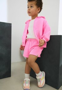 Veste à capuche rose vif et shorts assortis en tissu doux. Baskets avec des accents pastel et chaussettes blanches. Fond neutre.