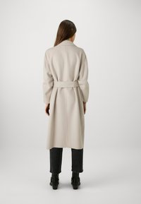 Cappotto lungo beige con cintura in vita, maniche larghe e una trama liscia. Indossato sopra jeans scuri e stivali neri alla caviglia.