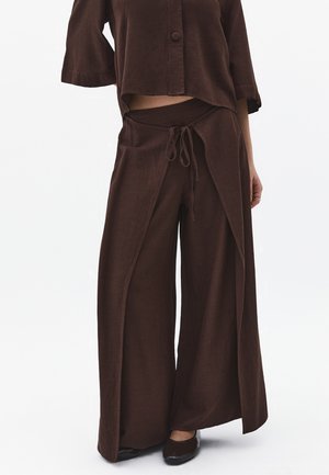 Kvinde iført mørkebrune vidde bukser med forreste overlægningspanel og matchende kort knapbluse mod hvid baggrund.