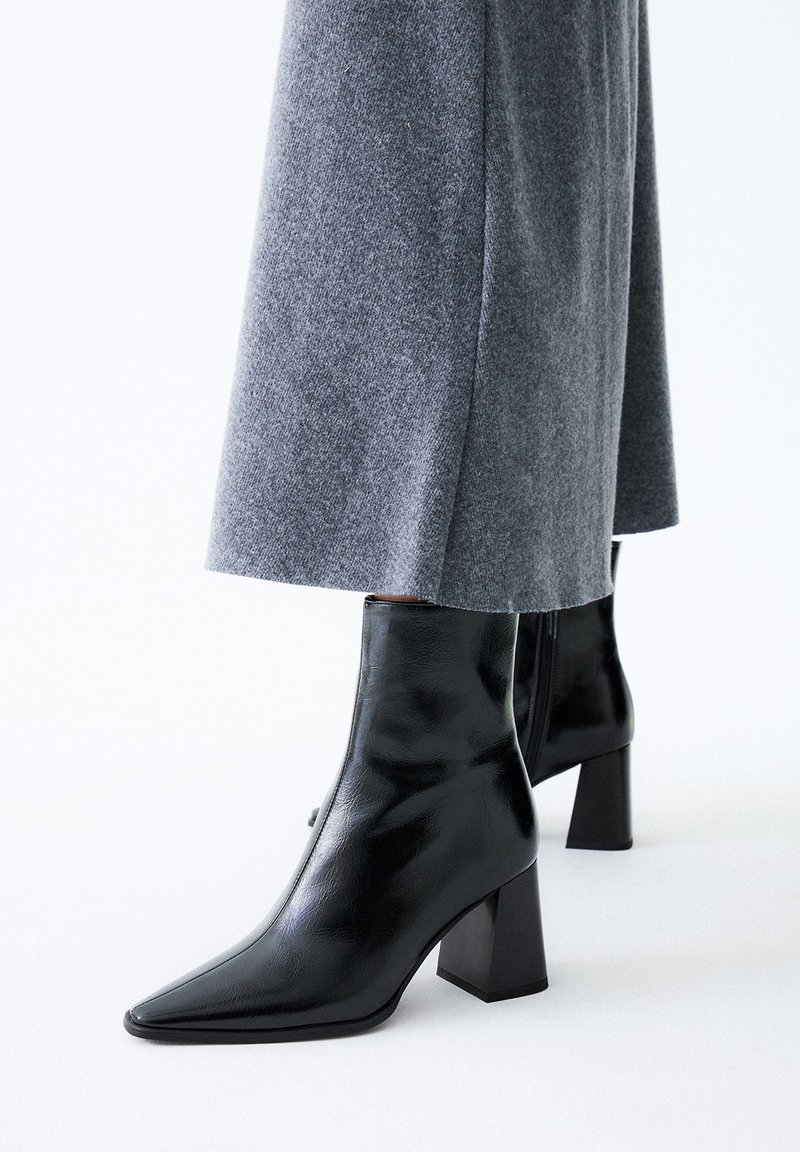 Botas de piel negra hasta el tobillo con punta afilada, tacón cuadrado y textura suave, combinadas con una falda de lana gris.
