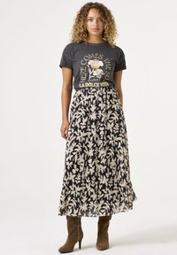 Grafisches T-Shirt in Anthrazitgrau mit Blumenmuster, kombiniert mit einem schwarzen und cremefarbenen, plissierten Maxirock mit Blättermuster. Grüne Absatzstiefel.