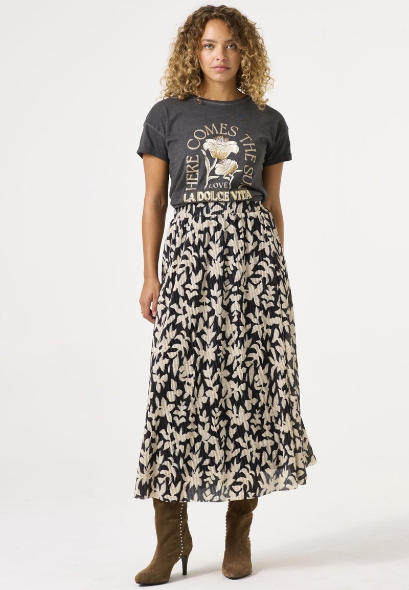 Camiseta gráfica en gris carbón con estampado floral, combinada con una falda maxi plisada en negro y crema con un patrón de hojas. Botines de tacón verdes.