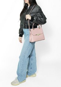 Borsa a mano rosa chiaro con dettagli dorati, design strutturato e manico superiore. Indossata con un bomber in pelle nera e jeans a zampa di elefante blu chiaro.