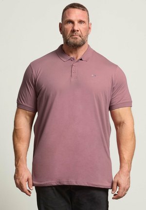 Skægget muskuløs mand med kort hår iført en mauve poloshirt og sorte bukser, stående mod en ensfarvet lys baggrund.