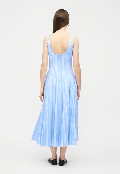 Γυναίκα που φοράει ένα αμάνικο midi φόρεμα σε ανοιχτό μπλε και άσπρες ρίγες με χαμηλή στρογγυλή πλάτη, μαύρα επίπεδα παπούτσια και κρίκους στα αυτιά, στέκεται με την πλάτη προς τα εμπρός.