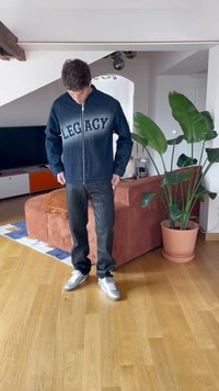 Tumši denima jaci ar uzrakstu "LEGACY" uz krūtīm, apvienota ar melniem biksēm un sudraba kedām. Iekštelpu vide ar augiem.