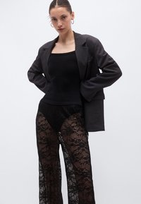 Must musta keha kujundav ülaosa koos tumeda bleiseriga ja läbipaistvate mustade pitspükstega, millel on lillemustriga, näidates kihilist ja tekstuurset välimust.