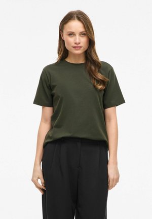 Frau mit welligem braunem Haar, die ein dunkelgrünes kurzärmeliges T-Shirt und schwarze hochtaillierte Hose vor weißem Hintergrund trägt.