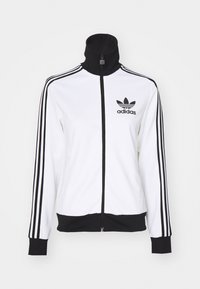 Giacca bianca con zip e colletto alto, dettagli neri e tre strisce orizzontali nere su ciascun manica. Presenta il logo Adidas nero sul petto.