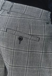 Szare spodnie w kratę wykonane z teksturowanej tkaniny, z tylną kieszenią z zapięciem na guzik. Charakteryzują się wyraźnym wzorem w kratkę.