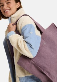 Fioletowa torba z korduroyu z paskami na ramię i małym detalem logo. Połączona z kremową i niebieską kurtką polarową o żebrowanej fakturze.