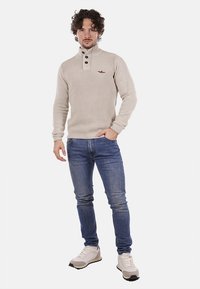 Maglione beige a costine con colletto alto, tre bottoni e un piccolo logo. Abbinato a jeans blu e sneakers bianche.