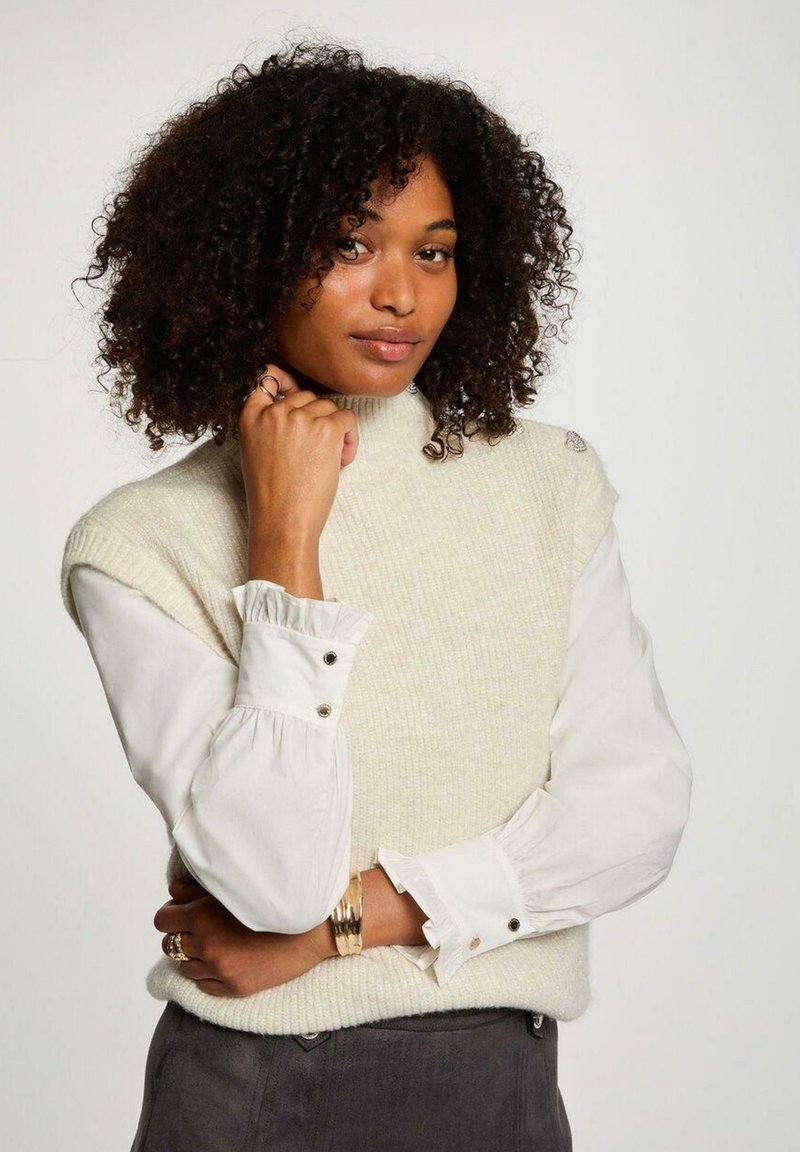 Maglione senza maniche in maglia crema sopra una camicia bianca con bottoni e polsini insellati. Presenta una finitura testurizzata e un semplice scollo rotondo.