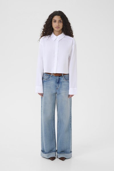 Chemise blanche courte à boutons avec manches longues, associée à un jean bleu clair à jambes larges, ceinturée d'une ceinture marron et portée avec des bottines marron à talons.