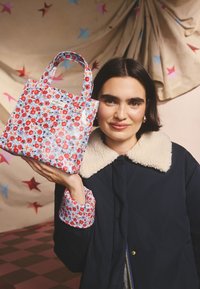 Sötét hajú nő aranykarika fülbevalóval, egy kis virágos táskát tartva, sötétkék kabátot visel, krémszínű sherpa gallérral.