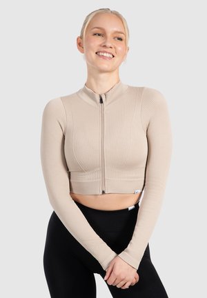 Beige gerippte Cropped-Jacke mit langen Ärmeln, hohem Kragen und Reißverschluss-Details an der Vorderseite, kombiniert mit schwarzen hoch geschnittenen Leggings.