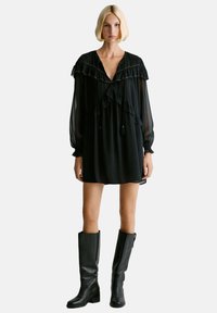 Mujer con un vestido negro holgado con detalles de volantes y mangas largas transparentes, combinado con botas de piel negras hasta la rodilla, de pie frente a un fondo blanco.