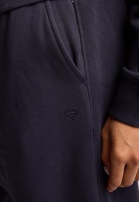 Felpa navy in un tessuto morbido e dalla texture fine. Presenta polsini a coste e un logo ricamato sulla tasca. Tasca laterale visibile.