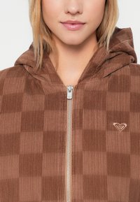 Donna che indossa una giacca con zip in velluto a coste marrone, con un piccolo logo a cuore ricamato sul petto.