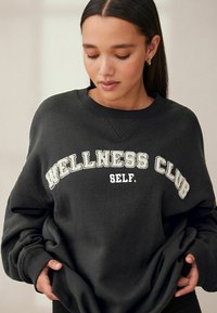 Musthaar mustrine, millel pehme kangas, millel on kontrastse valge tekstiga "WELLNESS CLUB" ja "SELF." Ribidega mansetid ja kaelus.