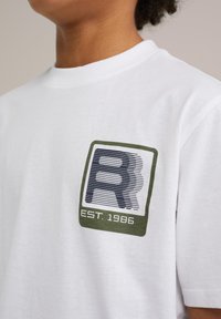 Balta medvilninė marškinė su žalia ir tamsiai mėlyna grafine emblema, kurioje yra "R" ir "EST. 1986" stačiakampio formoje ant krūtinės. Trumpi rankoviai.