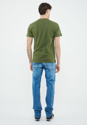 Uomo con tatuaggio floreale sul braccio, che indossa una maglietta verde oliva, jeans blu e sneakers nere, in piedi di spalle su sfondo bianco.