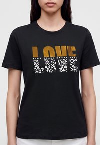 Γυναίκα που φοράει μαύρο T-shirt με τη λέξη LOVE σε χρυσό και λευκό leopard print, με μικρότερες λέξεις kind heart brave soul πάνω από τα γράμματα.