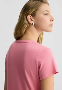 Rosa T-Shirt mit kurzen Ärmeln, runder Halslinie, weichem Material und lockerer Passform, kombiniert mit silbernen, herzförmigen Ohrringen.